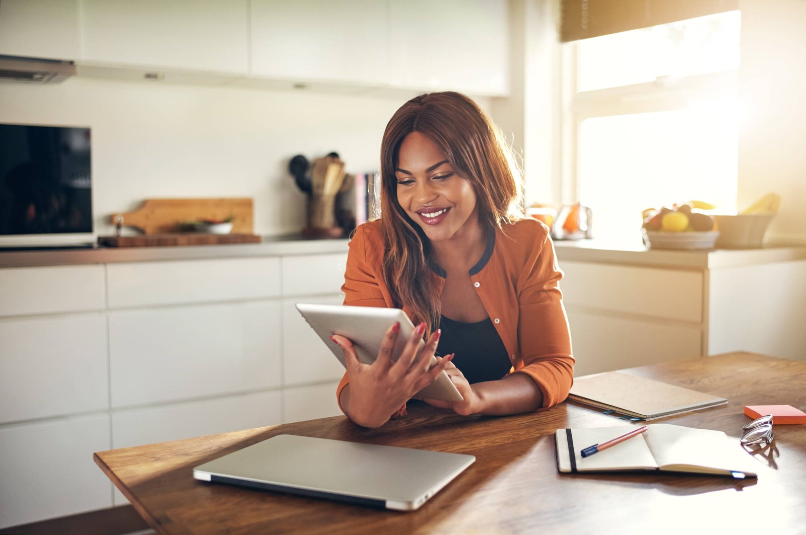 Smiling young African female entrepreneur working online with a digital tablet while sitting at a table in her kitchen